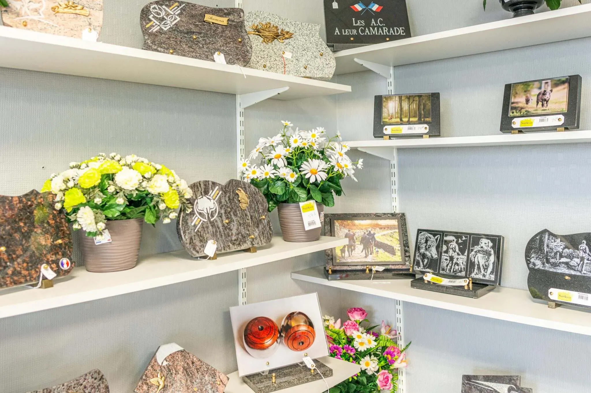 Rayonnages exposant des plaques funéraires gravées, des urnes et des pots de fleurs.
