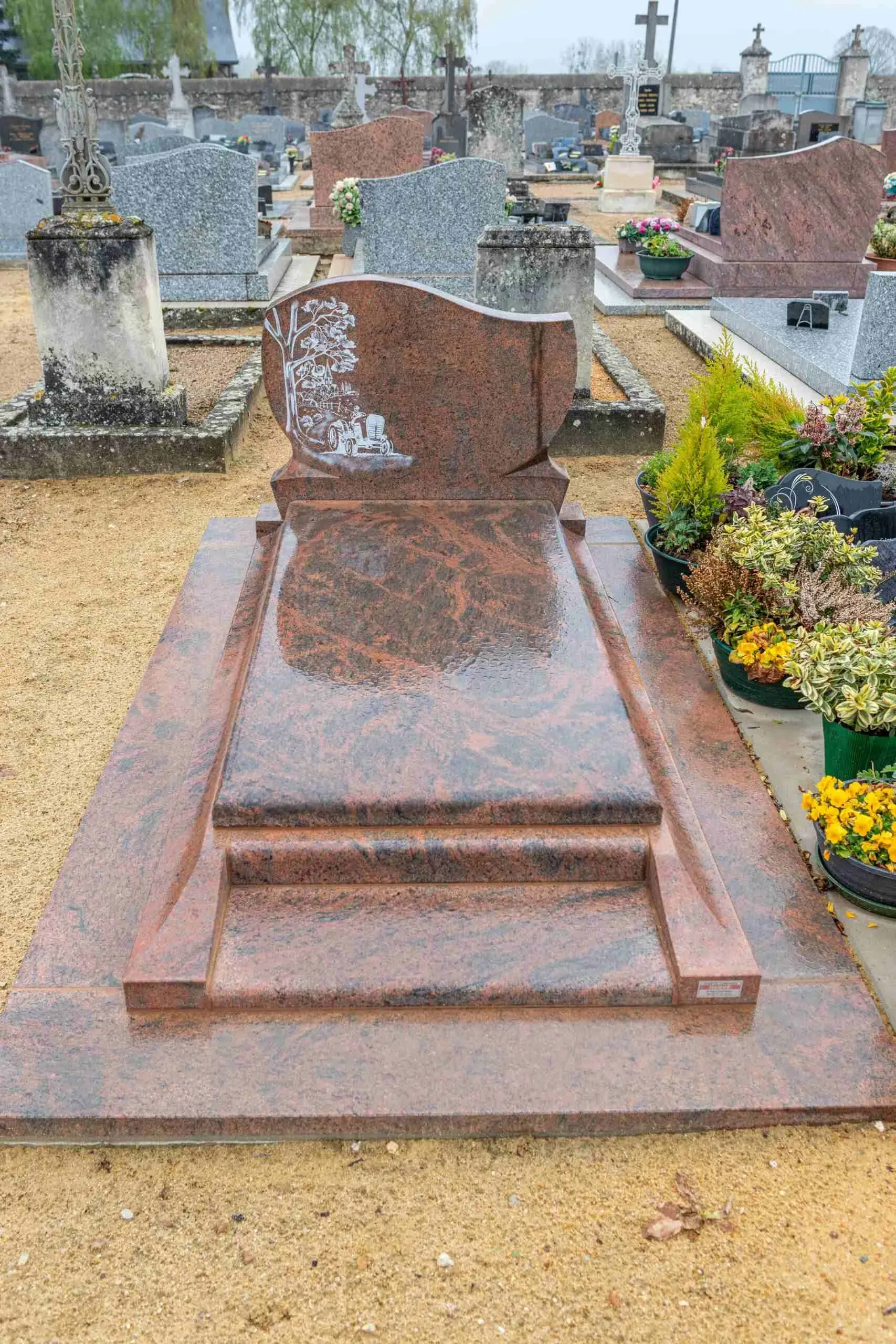 Tombale en granite rose veiné gravée d'un arbre et d'un tracteur dans un cimetière.