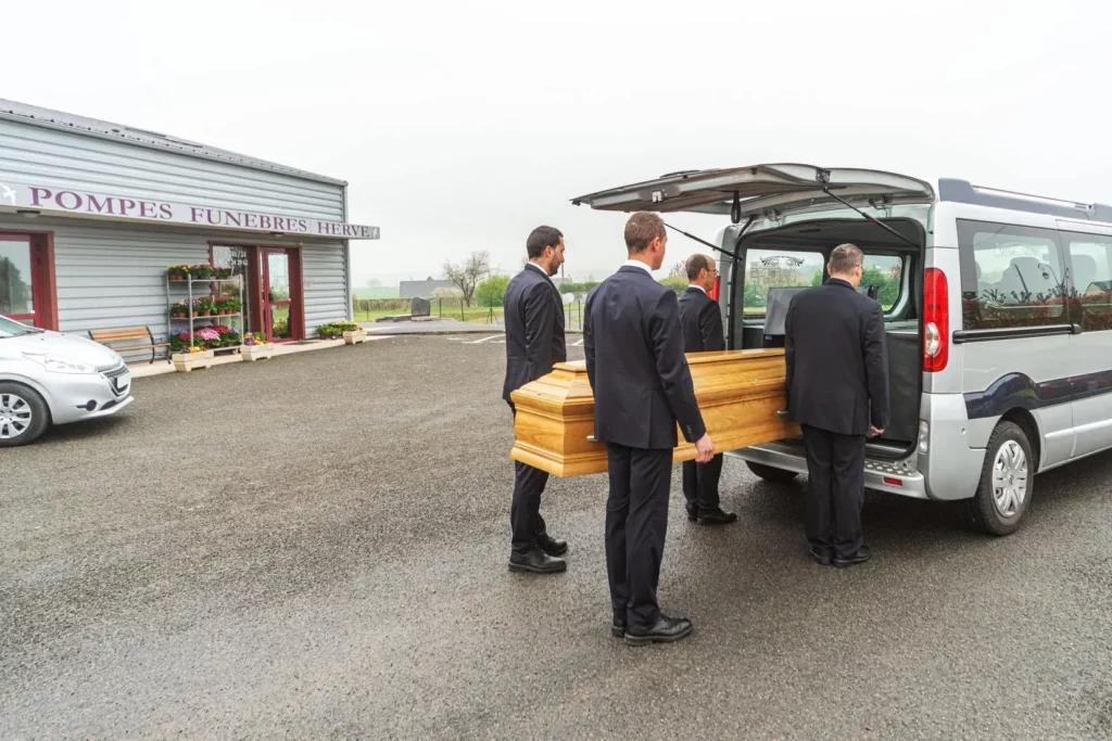 Quatre porteurs chargent avec respect un cercueil en bois dans le corbillard.